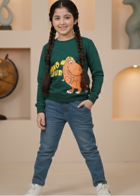 Girl wearing a green sweater with a dinosaur graphic and blue jeans indoors.