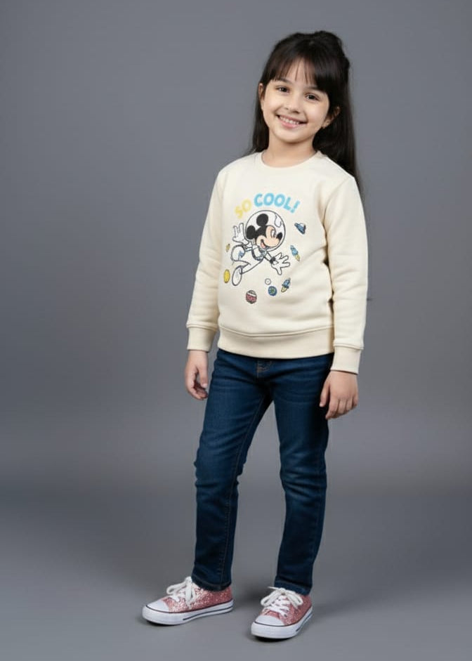 Young girl wearing a cream sweater with a cartoon character and dark jeans on a gray background