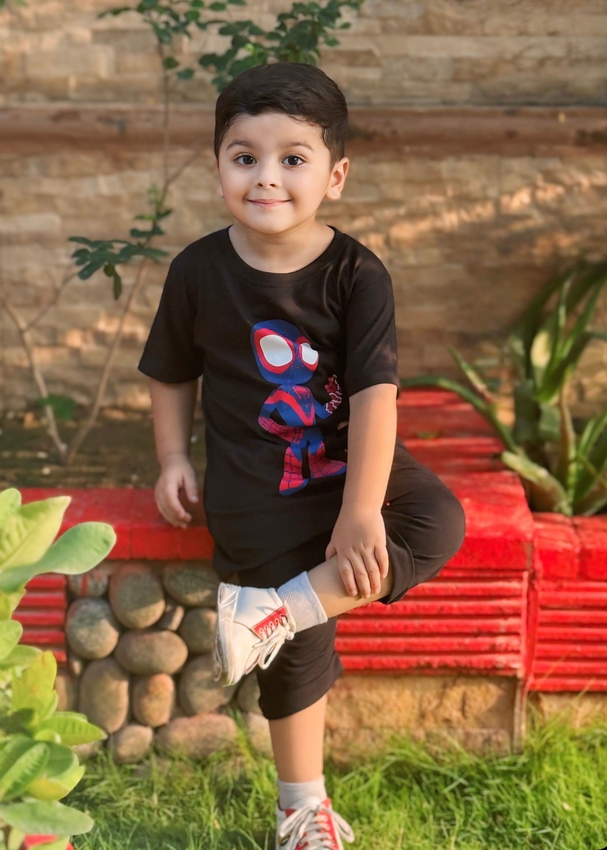 Child wearing a black t-shirt with a superhero design, standing outdoors with plants and a red structure in the background.