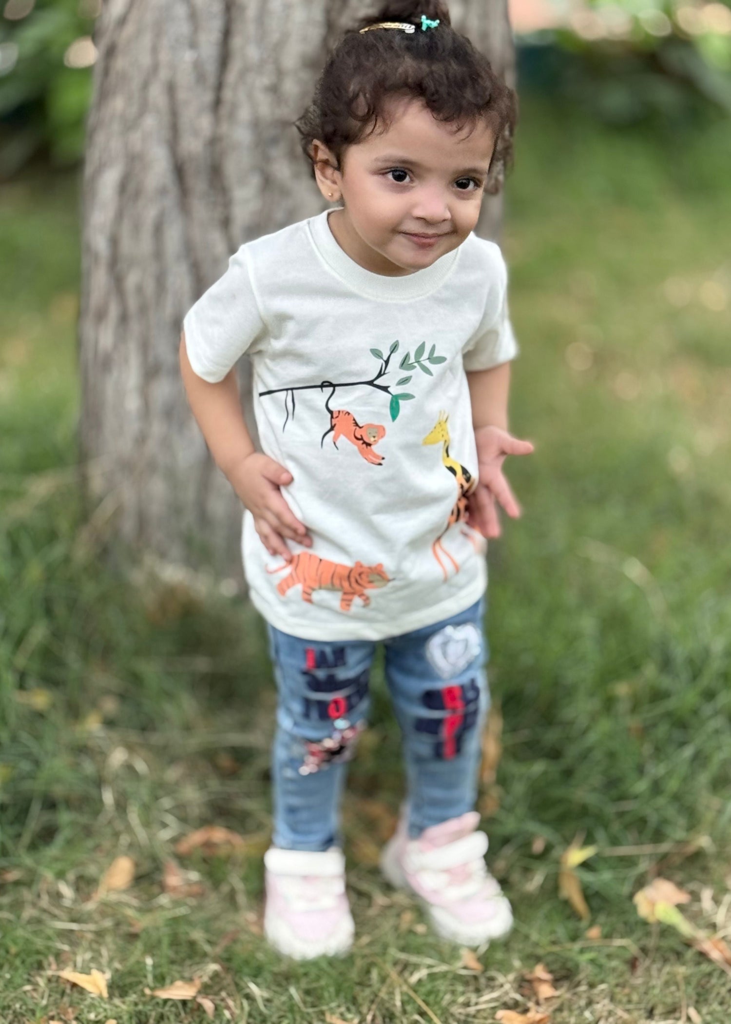 Child wearing a white shirt with animal prints and blue jeans standing outdoors.