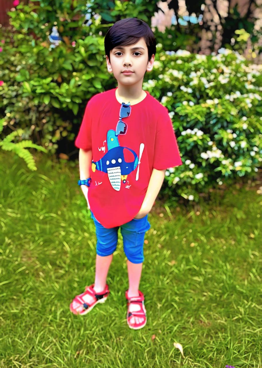 Child wearing a red shirt with a blue graphic and blue shorts standing on grass.
