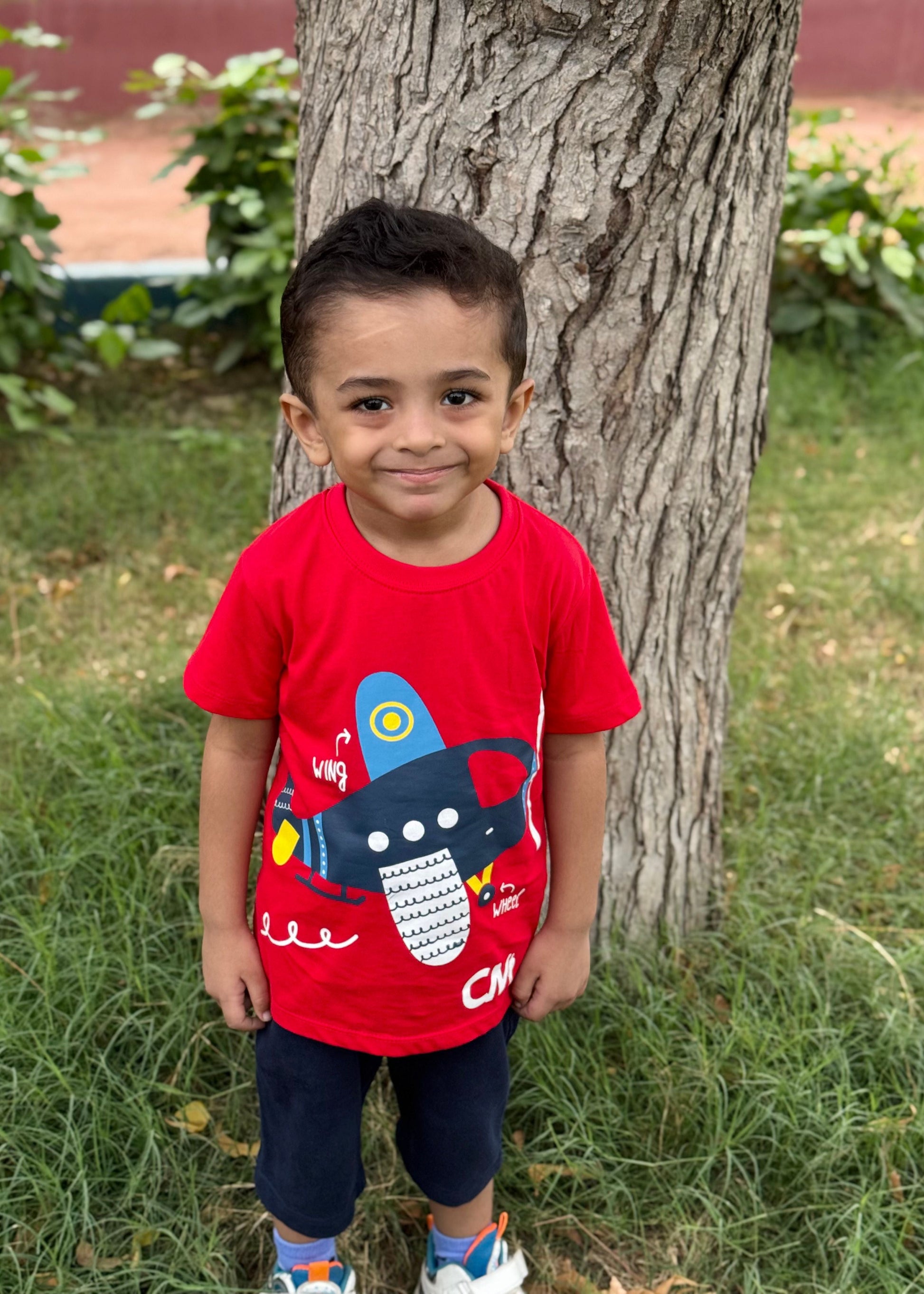Child wearing a red shirt with a cartoon character, standing next to a tree in an outdoor setting.