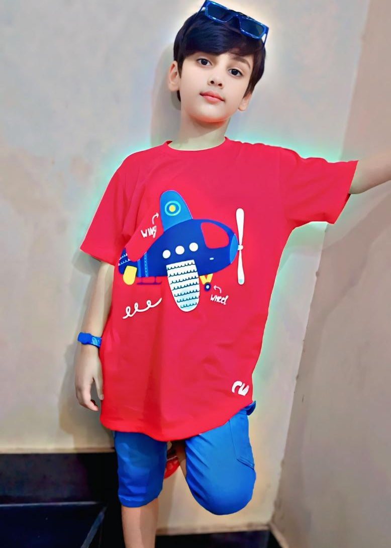Child wearing a red t-shirt with a colorful design and blue shorts indoors.