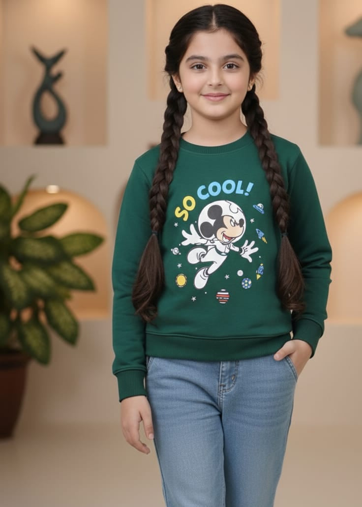 Young girl wearing a green sweatshirt with a graphic design and text, standing indoors.