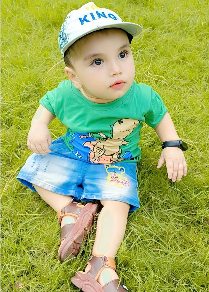 Child wearing a green shirt with a dinosaur design and blue shorts, sitting on grass.