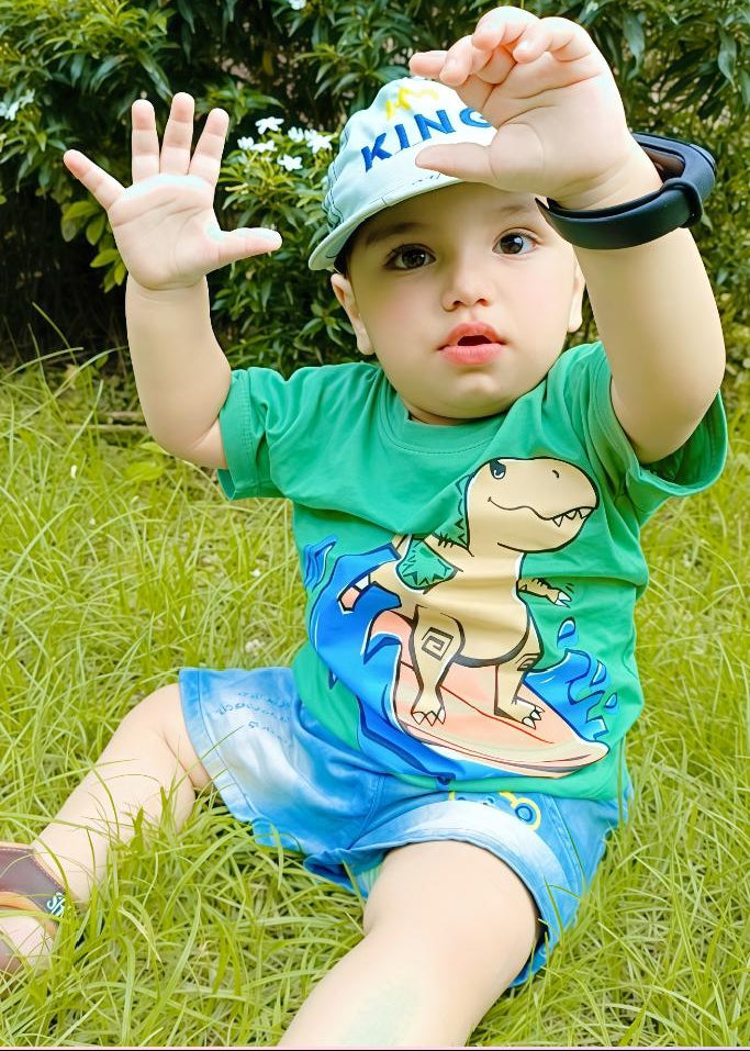 Child wearing a green shirt with a dinosaur design, sitting on grass.
