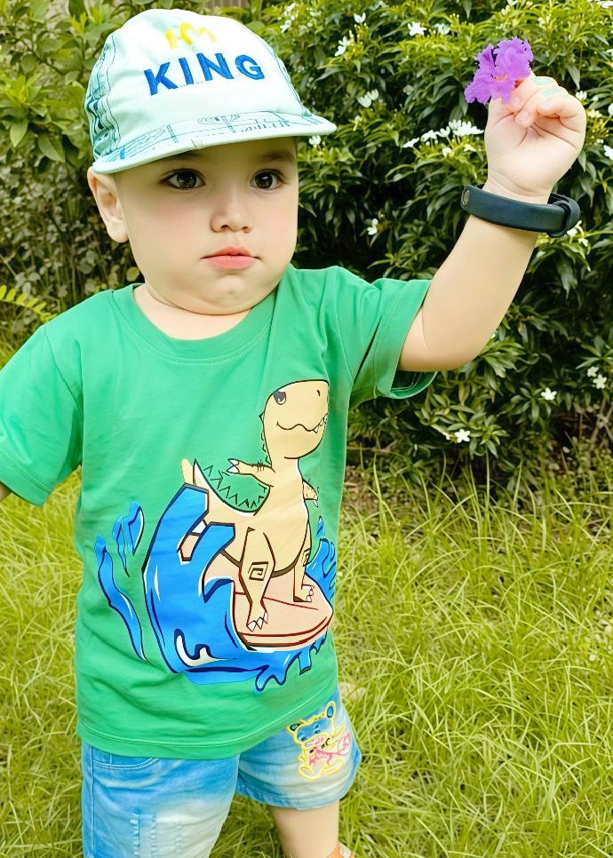 Child wearing a green shirt with a dinosaur design, blue shorts, and a white cap with 'KING' text outdoors.