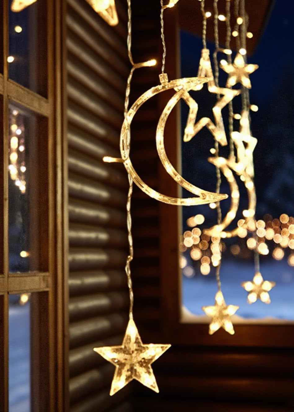 Decorative string lights with moon and star designs hanging by a window at night.