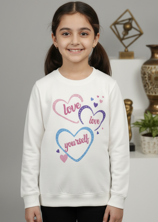 Young girl wearing a white sweatshirt with colorful text and heart designs, standing against a neutral background.
