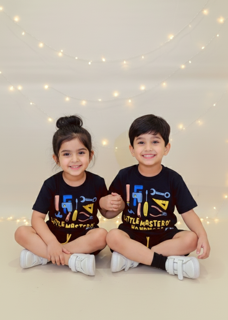 Two children wearing matching black t-shirts with colorful designs, sitting on a light-colored floor with string lights in the background.